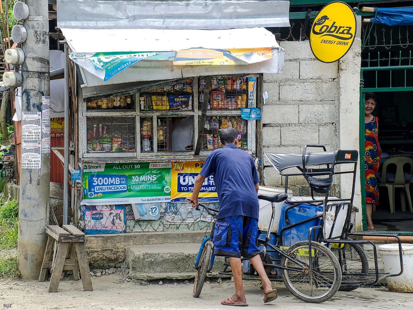 Daytime corner store with cola fascia and noodle tarp beside a jeepney.